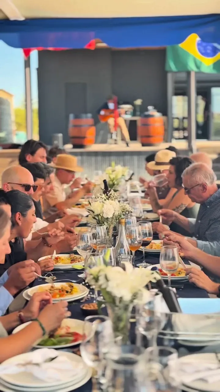 Invitados a la mesa larga bajo la carpa de Rhenus, con copas de vino naranjo y flores blancas; al fondo, un músico con guitarra entre dos barricas y la bandera de Brasil